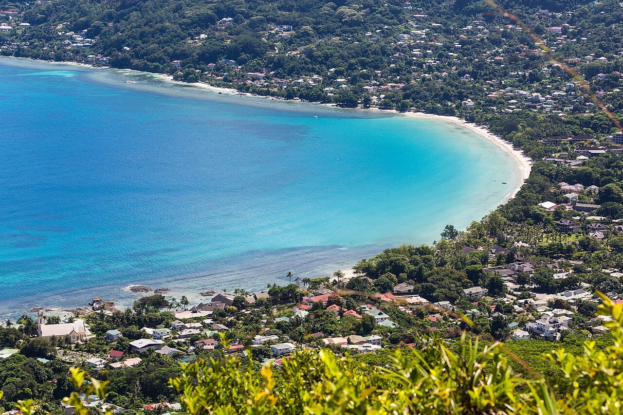 Ilmakuva rinteeltä Beau Vallonin lahden yli, talot ja puut edessä, pitkä kaartuva hiekkaranta ja turkoosi matalikko